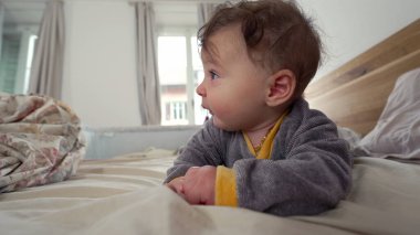 Bebek yatakta yüzüstü yatar, kameraya doğrudan meraklı ve meşgul bir ifadeyle bakar, erken motor beceri geliştirme, sıcak aile ortamı, keşif ve büyüme