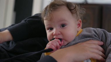 Bebek anne kolunda yatarken siyah kayışı çiğniyor, odaklanmış ve meraklı ifade, ağzıyla keşfetmek, kapalı alanda eğlenceli bir aile anı, dokunsal algısal keşif