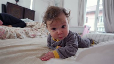 Bebek yatakta yüz üstü yatıyor, kameraya tuhaf bir ifadeyle bakıyor, göbek aktivitesiyle meşgul, erken motor beceri gelişimi, ev içi aile ortamları