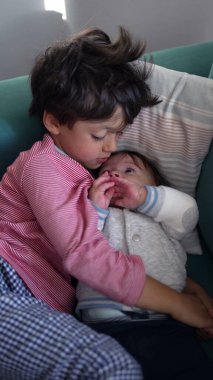 Kanepede bebeğe sarılan genç çocuk, hassas kardeş bağı, koruyucu kucaklama, huzurlu dinlenme, içten aile bağı, gerçek aşk ve rahat ev ortamında sıcaklık.