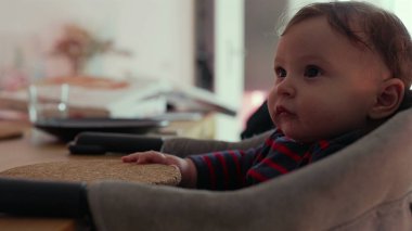 Bebek yüksek sandalyede oturuyor, bir testere tutuyor ve garip bir ifadeyle yalıyor, duyusal etkileşim yoluyla dokuyu keşfediyor, erken gelişim evresi, kapalı bir aile ortamı.