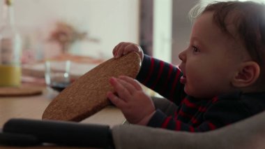 Bebek yüksek sandalyede oturuyor, bir testere tutuyor ve garip bir ifadeyle yalıyor, duyusal etkileşim yoluyla dokuyu keşfediyor, erken gelişim evresi, kapalı bir aile ortamı.