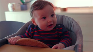 Bebek yüksek sandalyede oturuyor, tuhaf bir ifadeyle yan tarafa bakıyor, ağzını açıyor, heyecan gösteriyor, sıcak aile anı, erken sosyal gelişim ve nişan.