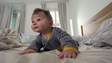 Bebek yatakta yüzüstü yatıyor, meraklı ve oyuncu bir ifadeyle ileri uzanıyor, erken motor beceri geliştirme, kapalı bir aile ortamı, erken çocuklukta keşif ve büyüme