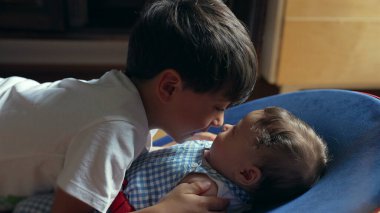 Küçük çocuk küçük kardeşlerin burnuna hafifçe dokunuyor, yumuşak bir bağ kuruyor, kardeş sevgisi, çocukluk bağı, hassas aile etkileşimi, samimi ve sevgi dolu bir sahne.