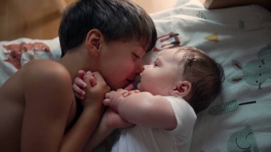 Küçük çocuk yatakta yatarken küçük kardeşine bakıyor. Yumuşak bir bağ anı, çocukluk bağı, aile sevgisi, kardeş ilişkisi, barışçıl ve sevecen bir ortam.