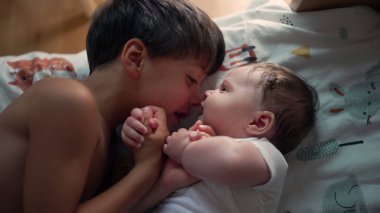 Küçük çocuk yatakta yatarken küçük kardeşine bakıyor. Yumuşak bir bağ anı, çocukluk bağı, aile sevgisi, kardeş ilişkisi, barışçıl ve sevecen bir ortam.