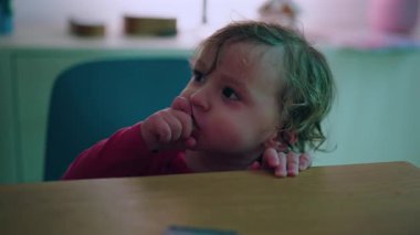 Little child holding food and chewing while standing behind table, candid family moment with messy face and innocent look