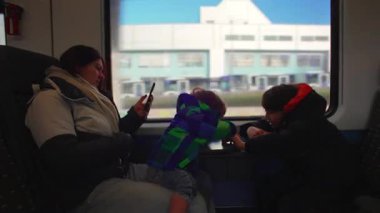 Family shares calm train ride, mother and two boys seated together while countryside passes outside window, evoking connection, travel routine, and childhood curiosity