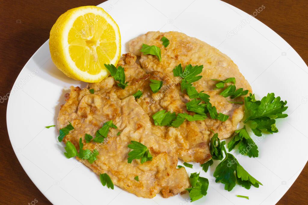 Pork escalopes in a white plate with half a lemon and parsley on a wooden table.