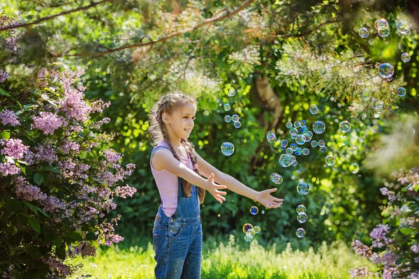 Chica encantadora en overoles de mezclilla la captura de burbujas de jabón en el jardín de color lila en flor — Foto de Stock