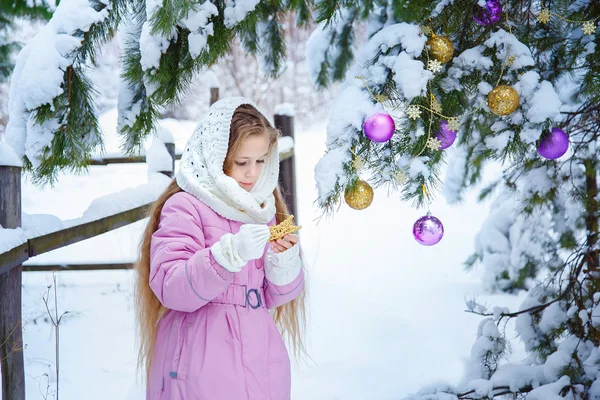 A cute little girl in a pink coat and white scarf adorns a Christmas pine tree in a snowy forest — Stock Photo, Image