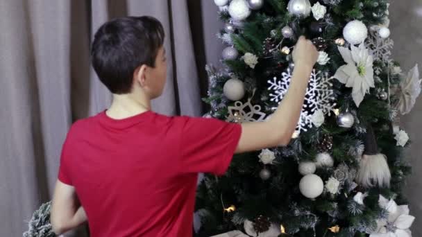 Мальчик-подросток украшает елку белыми игрушками — стоковое видео