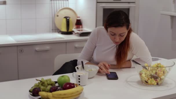 Chica se sienta a la mesa con teléfono comer ensalada de frutas — Vídeos de Stock