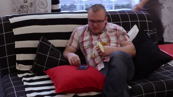 Man on couch eating banana and using phone — Stock Video