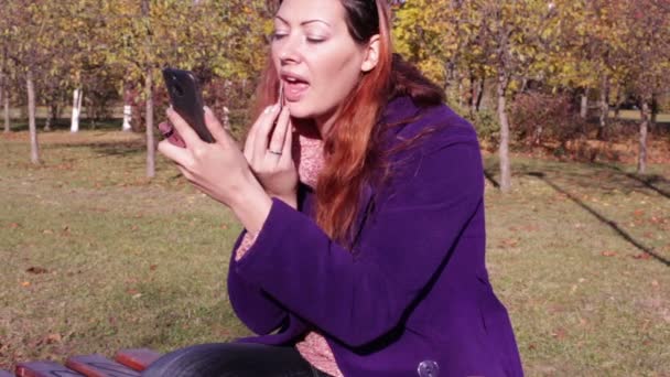Жінка малює губи, дивлячись у дзеркало по телефону — стокове відео