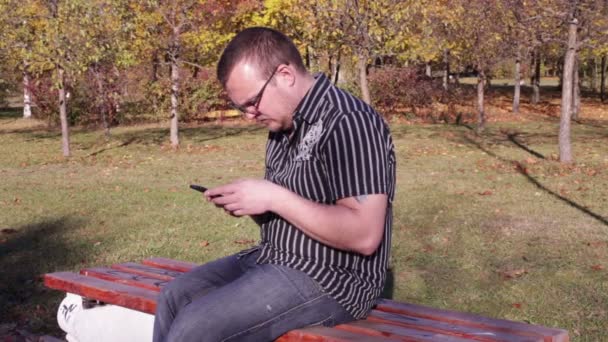 L'uomo si siede su una panchina del parco e digita un sms sul telefono — Video Stock