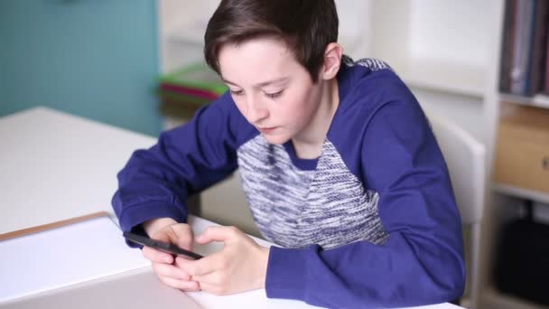 Підліток сидить за столом, граючи на вашому телефоні — стокове відео