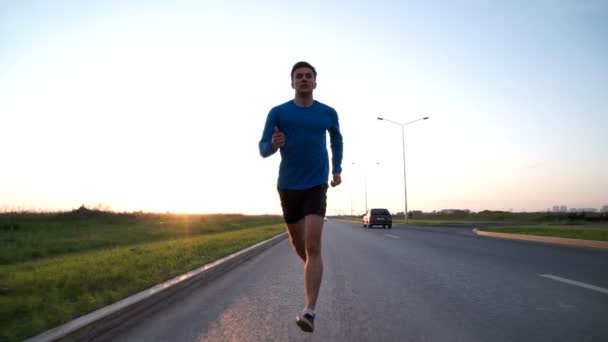 El tipo corre por la carretera al atardecer. Vista frontal — Vídeos de Stock