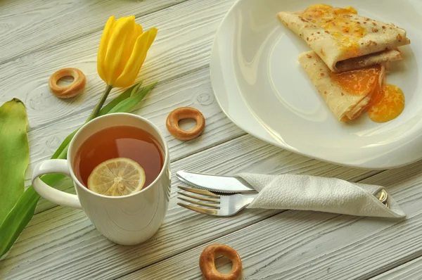 Аппетитные блины с фруктовым вареньем, чашка чая с лимоном, желтые тюльпаны, вилка и нож на деревенской белой деревянной поверхности — стоковое фото