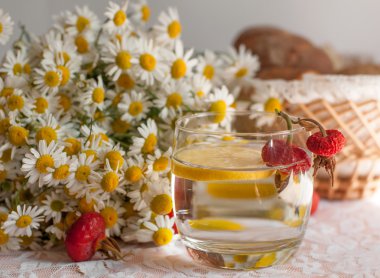 İçinde limon dilimi olan bir bardak su, bir buket chamomiles ve kalçalarla süslenmiş açık dantel yüzeyde bir tabak olgun erik