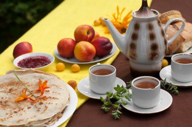 Çay, çay potu, bir tabak krep, pasta, olgun meyve ve alan çiçekleri parlak sarı ve kahverengi masa örtüsü