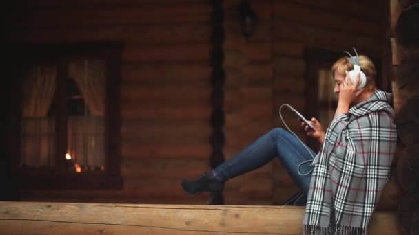 Fille avec le téléphone dans les écouteurs écoute de la musique sur la véranda en bois 