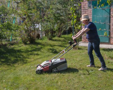 Baharda çalışan hasır şapkalı yaşlı bir kadın arka bahçesindeki çimleri biçiyor. Bahçe işleri, iş ve doğa kavramları. Yetişkinlikte hareket kavramı. Sezon bitmeden çim bakımı.