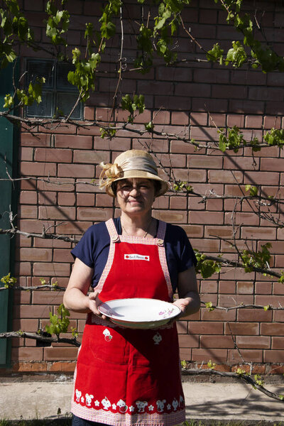 An elderly woman in a straw hat and red apron holds plate in her hands. Spring.