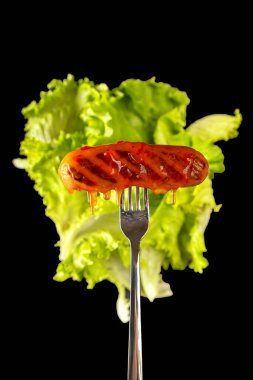 Çatalda ızgara sosis, arka planda marul yaprakları. Siyah bir arka planda izole edilmiş. Kolesterol, abur cubur. Seçici odak.