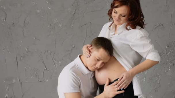 A handsome young man nuzzles the belly of his pregnant wife. Young wife pats him on the head and look at the camera. — Stock Video