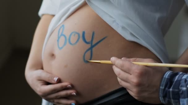 Ein junger kreativer Ehemann schrieb die blaue Farbe auf den Bauch des Wortes Junge. der Bauch einer glücklichen Schwangeren. — Stockvideo