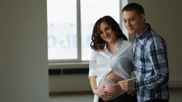 Молодий творчий чоловік написав синю фарбу на животі слова "хлопчик". Стоячи поруч із щасливою вагітною дружиною . — стокове відео