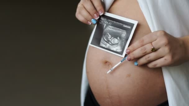 Mujer embarazada joven sosteniendo ultrasonido del abdomen. En su mano izquierda una prueba de embarazo . — Vídeos de Stock