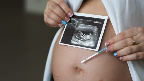Una mujer joven embarazada sostiene una imagen y una prueba de embarazo en el área abdominal . — Vídeos de Stock