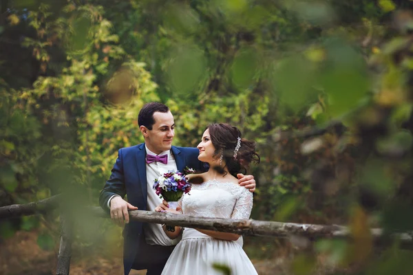 Bonito par de amantes recém-casados abraçando nas madeira.Mãos segurando um buquê de lilás branco lilás. Foto através das folhas das árvores . — Fotografia de Stock