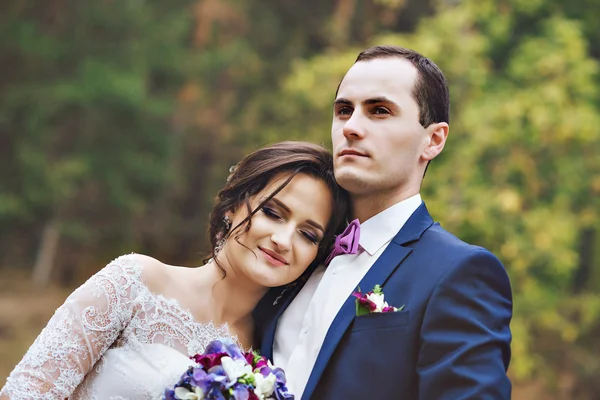 Hermosa pareja de amantes recién casados abrazándose en el bosque, una futura esposa en vestido de encaje, en el bosque. La novia se encuentra en el hombro del novio.. Vestido de novia blanco. Traje de hombre azul . —  Fotos de Stock