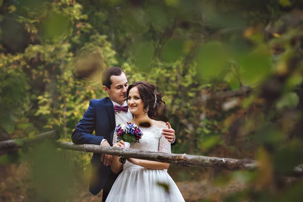 Hermoso par de amantes recién casados abrazándose en el bosque.Manos sosteniendo un ramo de lila blanca lila. Foto a través de las hojas de los árboles. Se ríen —  Fotos de Stock