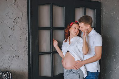 Neşeli genç adam ve güzel bir hamile kadının birbirlerine sarılma. Biz birbirimize bakıyoruz ve midsection sarıl. Fashion photoshoot stüdyoda. Kot pantolon ve beyaz gömlek bir kadında. 