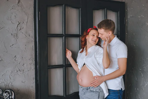 Neşeli genç adam ve güzel bir hamile kadının birbirlerine sarılma. Biz birbirimize bakıyoruz ve midsection sarıl. Fashion photoshoot stüdyoda. Kot pantolon ve beyaz gömlek bir kadında. 