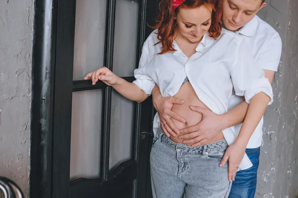 Joyful young man and a beautiful pregnant woman hugging each other. Laughing couple expecting a baby. Man embraces naked pregnant tummy. We look at each other and hug the midsection. — Stock Photo, Image