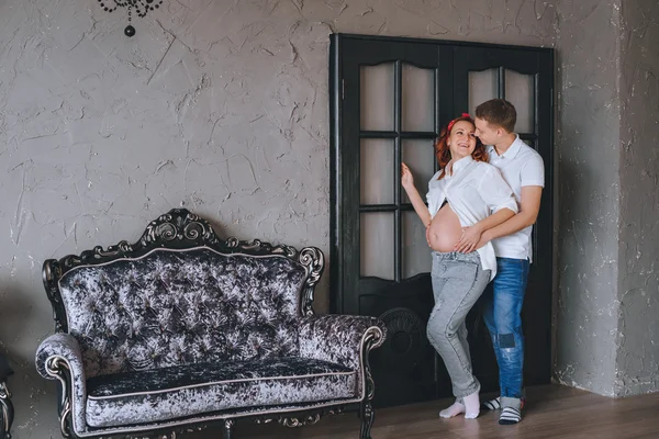 Joven alegre y una hermosa mujer embarazada abrazándose. Nos miramos el uno al otro y abrazamos la sección media. Sesión de fotos de moda en el estudio. Una mujer en jeans y una camisa blanca . — Foto de Stock