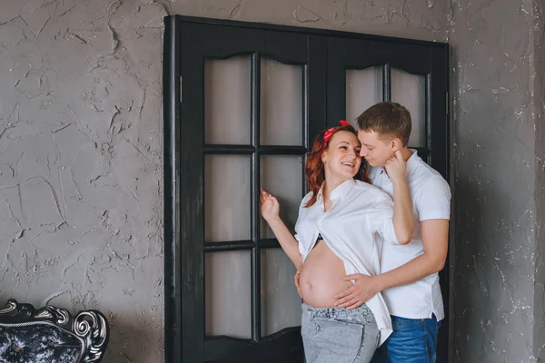 Joyeux jeune homme et une belle femme enceinte se serrant dans leurs bras. On se regarde et on embrasse la section médiane. Photoshoot de mode en studio. Une femme en jeans et une chemise blanche . — Photo