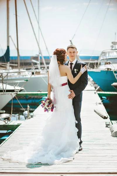 La pareja de pie en el muelle en el puerto, el novio abraza a la novia. Vestido de novia blanco con un tren . —  Fotos de Stock