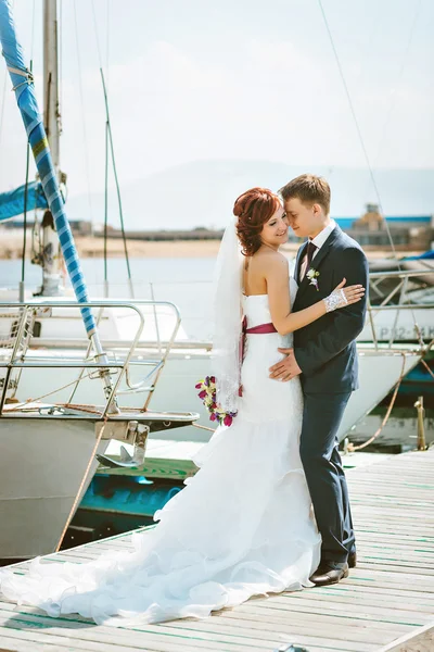 O casal que está no cais no porto, noivo abraça noiva. Vestido de noiva branco com um trem . — Fotografia de Stock