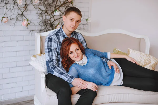 Pareja joven embarazada en el sofá del amor. la mujer yacía de rodillas de los hombres. marido abraza a su esposa. Con colores rosa cuarzo y azul cielo . — Foto de Stock