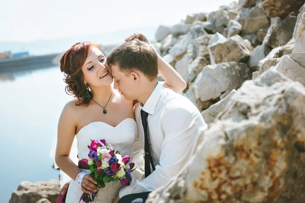 Lyckligt leende unga bruden och brudgummen, promenader på stenar, vacker himmel, vatten. Paret älskar varandra. — Stockfoto