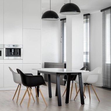 Modern masası ve iki siyah kolye lambası altında dört sandalyesi olan şık siyah-beyaz yemek salonu.