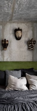 Duvarında ve tavanında çıplak beton olan modern yatak odasının dikey manzarası dekoratif Afrika tarzı maskeler ve yatak başlığında yeşil kadife.