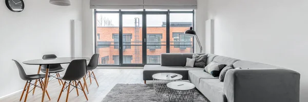 Panorama of modern apartment with window wall in stylish living room with dining area for four and big, gray corner sofa